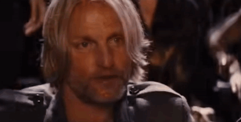 A Man With Long Blonde Hair And A Beard Is Sitting In A Dark Room With Other People . GIF