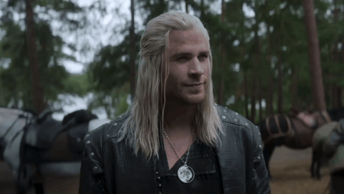 A Man With Long Blonde Hair And A Necklace Is Standing In The Woods . GIF