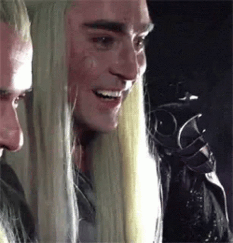 A Man With Long Blonde Hair Is Smiling And Looking At Another Man . GIF