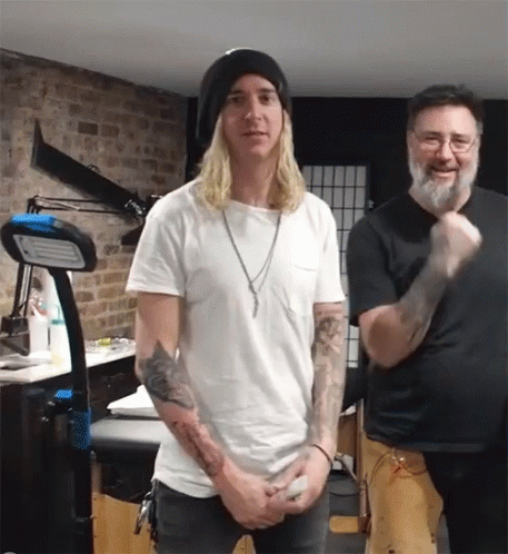 A Man With Long Blonde Hair Stands Next To Another Man With A Beard GIF