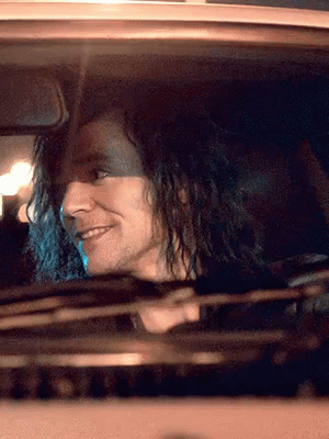 A Man With Long Curly Hair Is Driving A Car And Smiling GIF