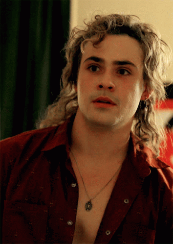 A Man With Long Curly Hair Is Wearing A Necklace And A Red Shirt GIF