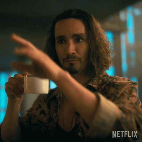 A Man With Long Hair And A Beard Is Holding A Cup Of Coffee And Pointing At It With A Netflix Logo In The Background GIF
