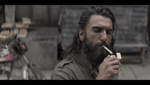 A Man With Long Hair And A Beard Is Lighting A Cigarette With A Lighter GIF