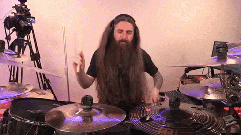 A Man With Long Hair And A Beard Is Playing Drums In Front Of A Camera GIF