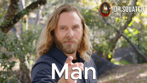 A Man With Long Hair And A Beard Is Pointing At The Camera And The Word Man Is On The Front Of His Shirt GIF