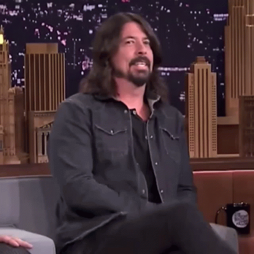 A Man With Long Hair And A Beard Is Sitting In A Chair And Waving His Hand GIF