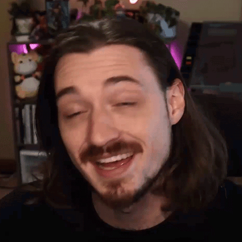 A Man With Long Hair And A Beard Is Smiling In Front Of A Bookshelf . GIF