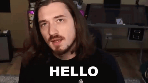 A Man With Long Hair And A Beard Is Standing In Front Of A Desk And Says `` Hello '' . GIF