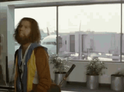 A Man With Long Hair And A Beard Is Standing In Front Of A Window Holding A Golf Club . GIF