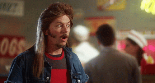 A Man With Long Hair And A Beard Stands In Front Of A Sign That Says Hot Dogs GIF