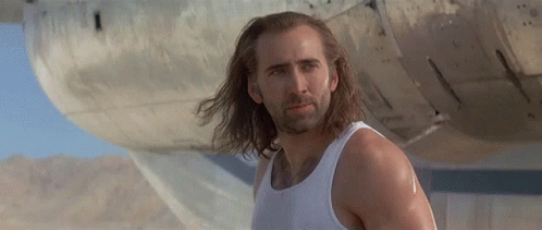 A Man With Long Hair And A Beard Wearing A White Tank Top Is Standing In Front Of A Plane . GIF