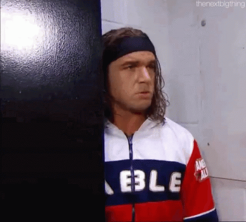 A Man With Long Hair And A Headband Is Leaning Against A Wall In A Room . GIF