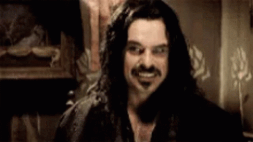 A Man With Long Hair And A Mustache Is Smiling And Looking At The Camera . GIF