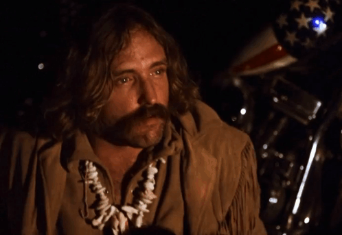 A Man With Long Hair And A Mustache Is Standing In Front Of A Motorcycle With An American Flag In The Background GIF