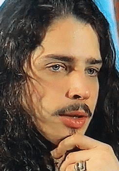 A Man With Long Hair And A Ring On His Finger Looks At The Camera GIF