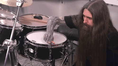 A Man With Long Hair And A Tattooed Arm Playing A Drum Set GIF