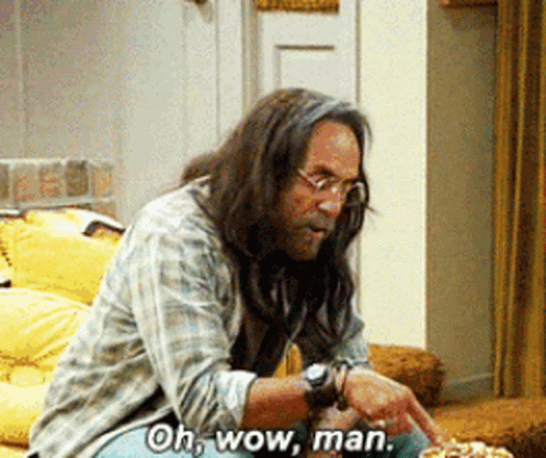A Man With Long Hair And Glasses Is Sitting On A Couch And Says Oh Wow Man GIF