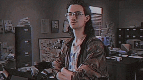 A Man With Long Hair And Glasses Is Standing In Front Of A Sign That Says Hackerman GIF