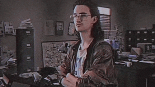A Man With Long Hair And Glasses Stands In Front Of A Sign That Says Hackerman GIF