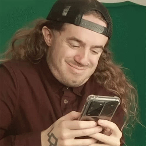 A Man With Long Hair Is Holding A Cell Phone In His Hands And Smiling . GIF