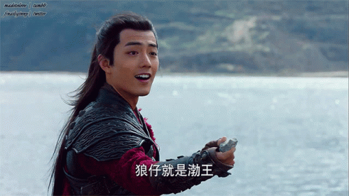 A Man With Long Hair Is Holding A Rock In Front Of A Body Of Water And Has Chinese Writing On His Arm GIF
