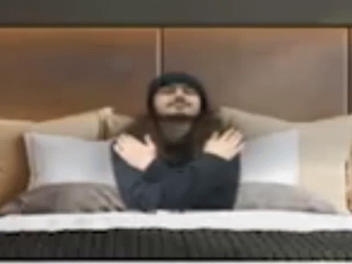 A Man With Long Hair Is Laying On A Bed With His Hands Folded . GIF