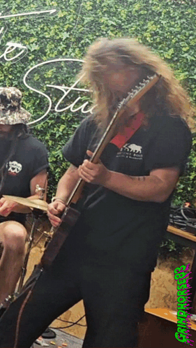 A Man With Long Hair Is Playing A Guitar In Front Of A Sign That Says Stu GIF