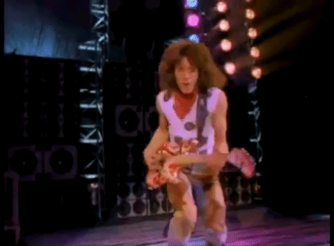 A Man With Long Hair Is Playing A Guitar On A Stage With Lights Behind Him . GIF