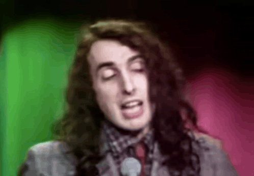 A Man With Long Hair Is Singing Into A Microphone In Front Of A Green And Pink Background . GIF