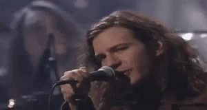 a man with long hair is singing into a microphone while another man looks on .