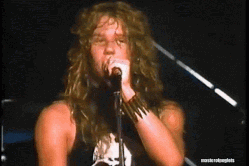 A Man With Long Hair Is Singing Into A Microphone While Standing On A Stage . GIF