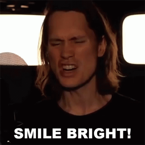 A Man With Long Hair Is Singing Into A Microphone With The Words Smile Bright Behind Him GIF