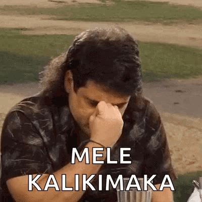 A Man With Long Hair Is Sitting At A Table With His Hand On His Forehead And The Words Mele Kalikimaka On The Screen . GIF