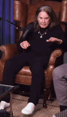 A Man With Long Hair Is Sitting In A Chair With His Feet Crossed GIF