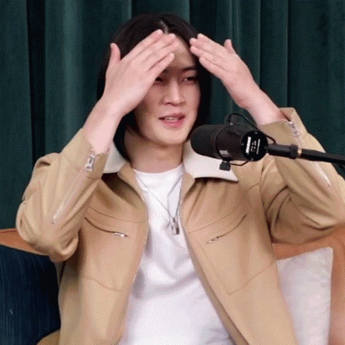 A Man With Long Hair Is Sitting In Front Of A Microphone And Covering His Head With His Hands . GIF