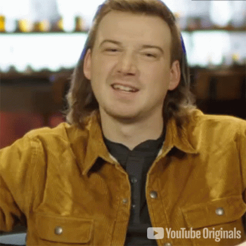 A Man With Long Hair Is Smiling And Wearing A Brown Jacket . GIF