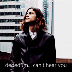 A Man With Long Hair Is Standing In Front Of A Building With The Words Dededum Can 'T Hear You Written Below Him GIF