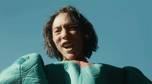 A Man With Long Hair Is Wearing A Green Blanket And Smiling GIF