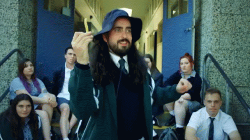 A Man With Long Hair Wearing A Blue Hat Stands In Front Of A Group Of Students GIF