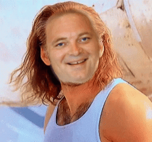 A Man With Long Red Hair Wearing A White Tank Top Smiles For The Camera GIF