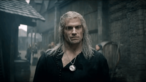 a man with long white hair and a necklace on his neck is standing in front of a brick building .