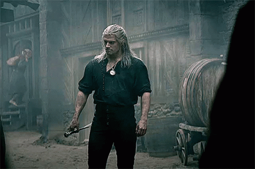 a man with long white hair is holding a sword in front of a building and barrels .