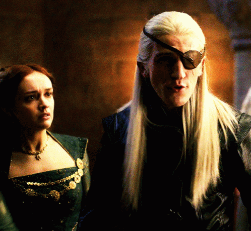 A Man With Long White Hair Wearing A Black Eye Patch Stands Next To A Woman GIF
