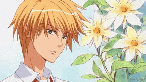 A Man With Orange Hair Is Standing In Front Of A Bush Of Flowers GIF