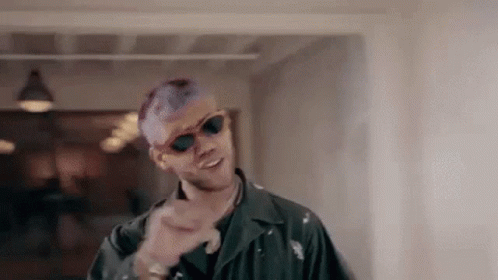 A Man With Purple Hair And Sunglasses Is Standing In A Hallway And Pointing At The Camera . GIF
