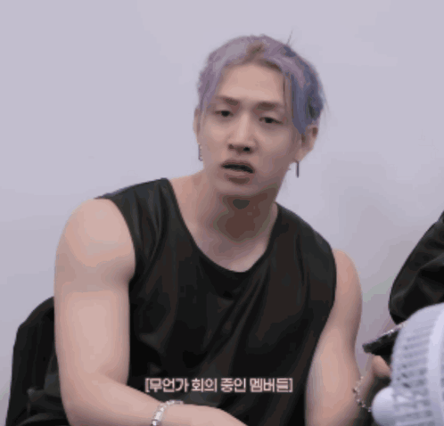 A Man With Purple Hair Is Wearing A Black Tank Top And Sitting Next To A Fan GIF