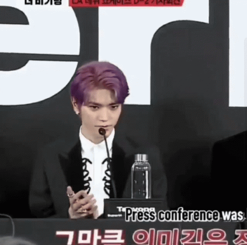 A Man With Purple Hair Speaking Into A Microphone In Front Of A Sign That Says Press Conference GIF