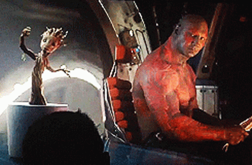 A Man With Red Paint On His Body Is Sitting In A Chair Next To A Statue Of Groot . GIF