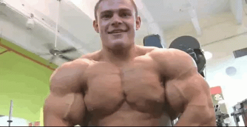A Man With Very Large Muscles Is Standing In A Gym And Smiling At The Camera . GIF